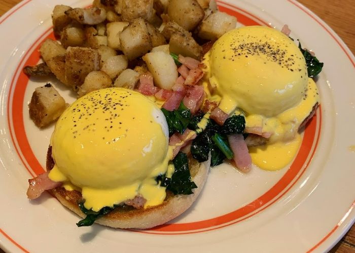 Eggs Benedict at Eggs ‘n Things ExpoCity, showing two poached eggs on top of spinach and bacon bits, served alongside potatoes and covered in sauce.